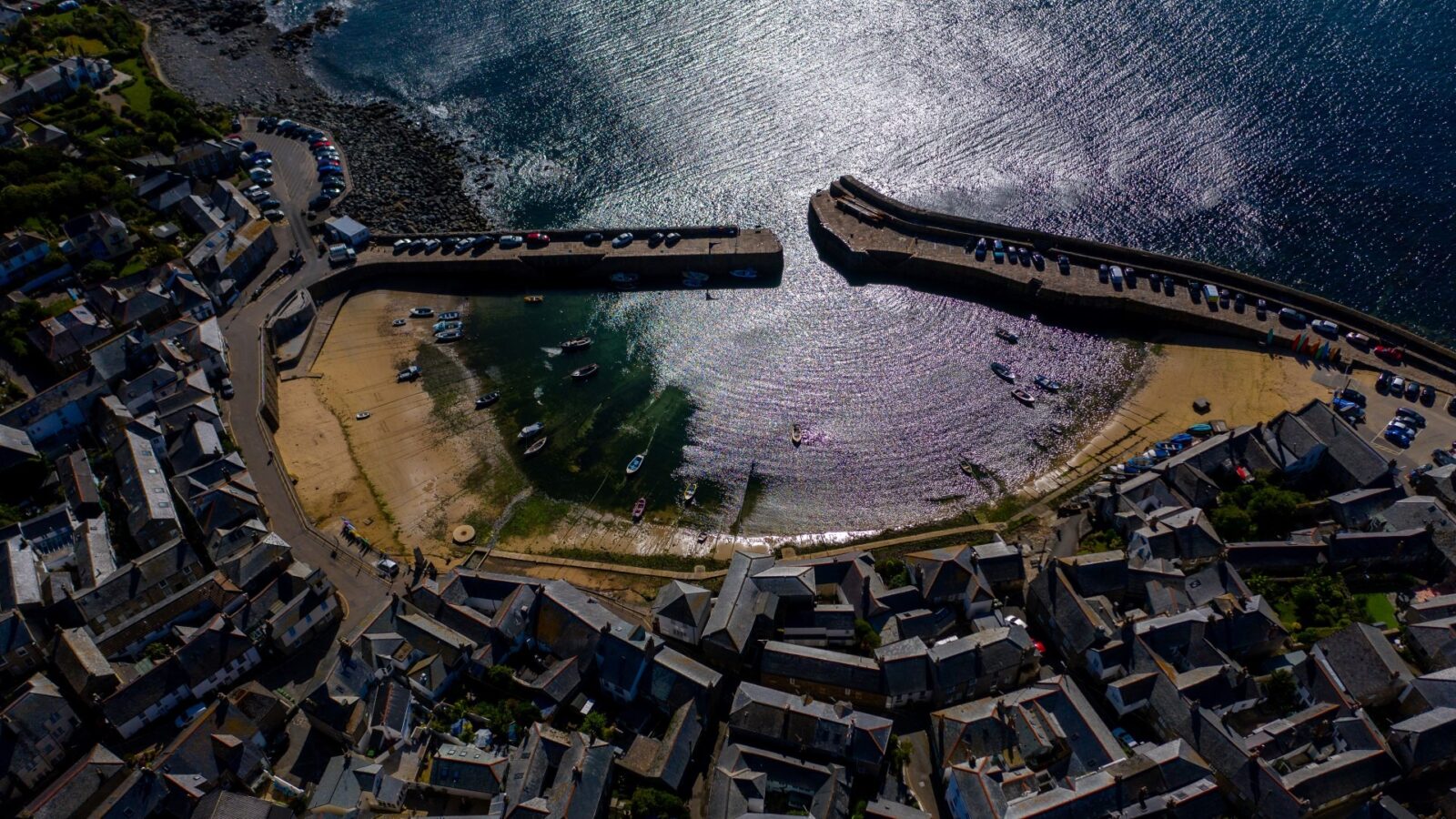 An aerial view of a quaint coastal village with a small harbor flanked by two curved piers resembles a hidden paradise. Sailboats and small vessels float in the shimmering water, like fallen angels at rest. Surrounding the harbor are densely packed houses with gray roofs, narrow streets, and a rocky shoreline.