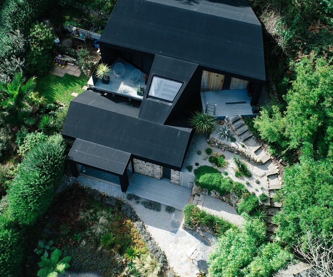 Aerial view of a modern, angular black house surrounded by lush greenery, like a fallen angel among the trees. The house features multiple levels and large windows. The front yard includes a well-designed garden with various plants, gravel pathways, and outdoor seating areas.