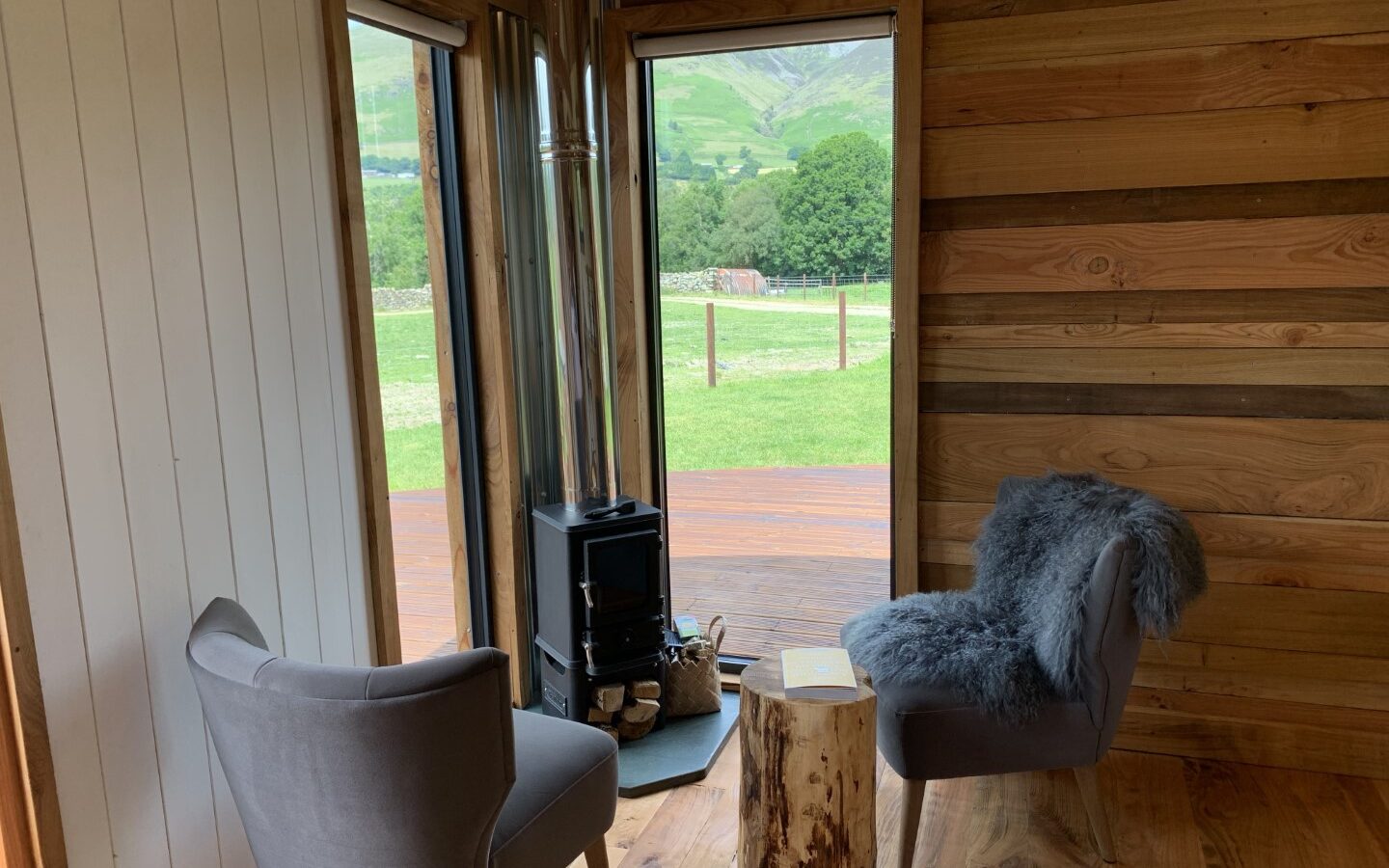 The cozy interior, featuring two gray chairs and a wood stove, frames a stunning window view of The Vale's lush green fields and majestic mountains.