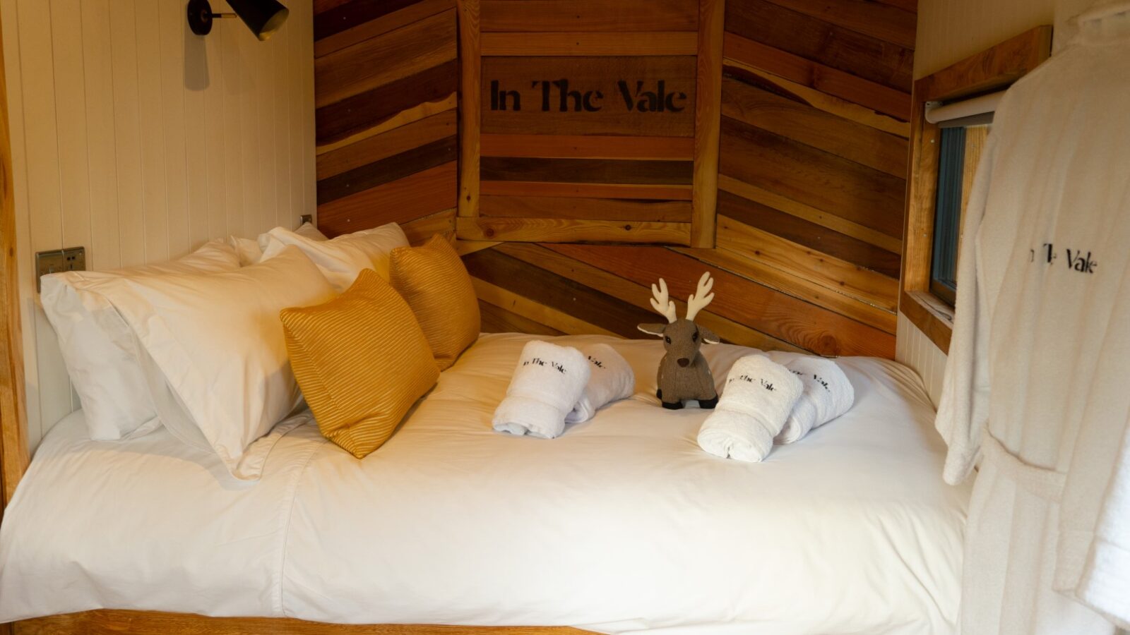 Cozy bed in The Vale with white linens and yellow pillows, featuring a plush deer and folded towels. A robe hangs on the wall.