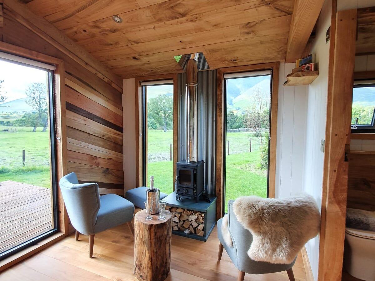 In the Vale's wooden interior, two chairs and a quaint log table complement a small wood stove, all overlooking a grassy landscape through the glass doors.