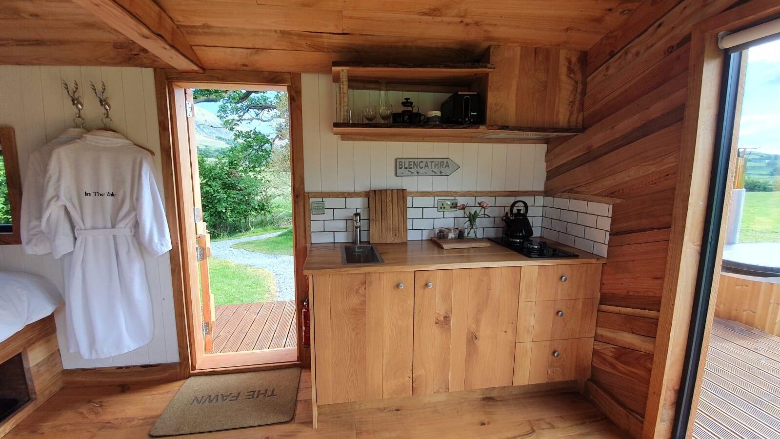 The Vale's cozy wooden cabin interior features a quaint kitchen, an inviting open door leading outside, and a robe hanging on the wall, embodying rustic charm.