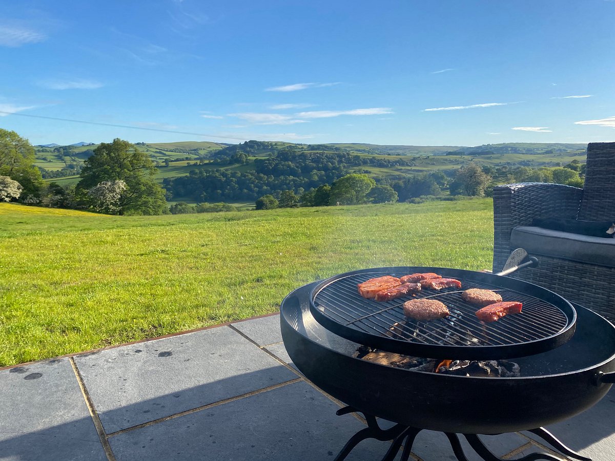 On the patio of Great House Farm, a barbecue grill sizzles with burgers and sausages, blending the luxury of glamping with the allure of a scenic landscape nestled among rolling hills and trees.