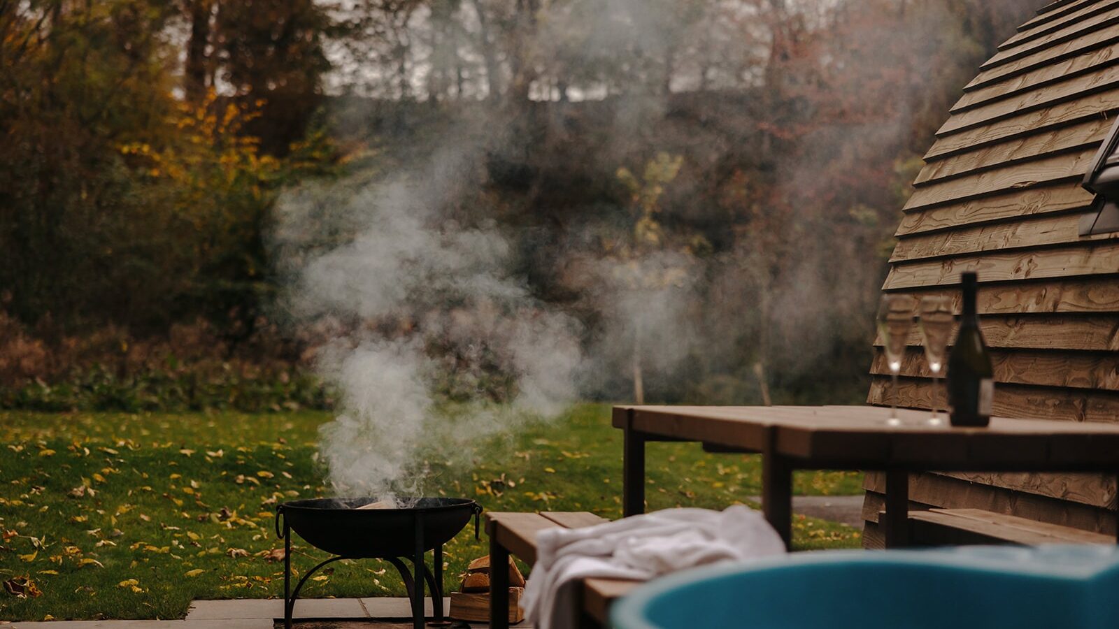 A steaming hot tub is in the foreground on a patio beside a wooden structure at Forcett Grange. Nearby, a grill lets off smoke, with a park-like landscape and trees with autumn foliage in the background. Towels and a bottle are placed on a wooden bench beside the hot tub.