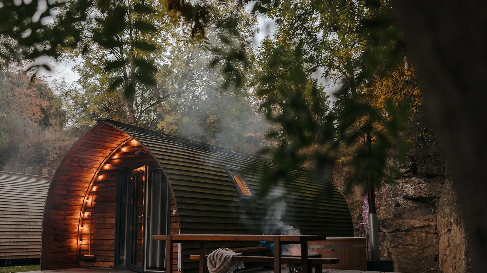 A cozy, wooden, pod-like cabin at Forcett Grange is nestled among trees, with soft exterior lighting illuminating the entrance. Smoke drifts up from a fire pit near a wooden picnic table in the foreground. The setting is serene and inviting, surrounded by autumn foliage.