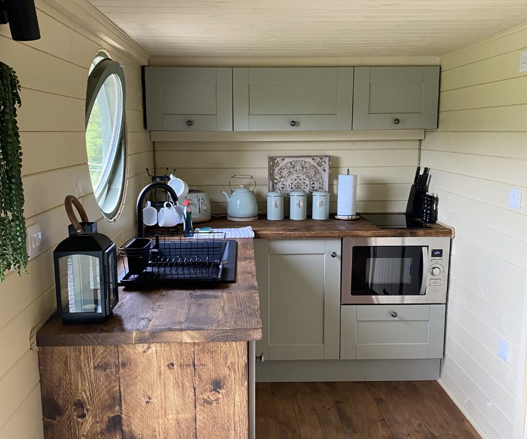 A small, cozy kitchen with light green cabinets, a wooden countertop, and wood-paneled walls and floor—perfect for a glamping retreat at Freedom Fields. It includes a microwave, coffee maker, kettle, and a dish rack. A circular window is on the far left wall. Decorative jars and kitchen utensils are neatly arranged.