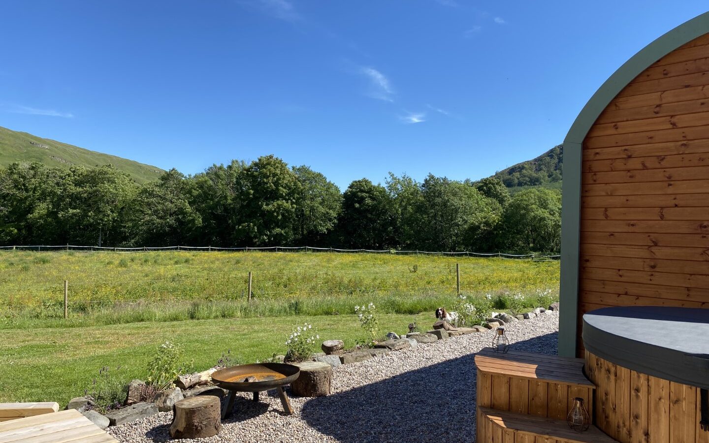 A scenic view of a lush green field bordered by trees under a clear blue sky. In the foreground, there is a small wooden deck with steps leading up to a wooden structure, perfect for glamping. A fire pit surrounded by tree stump seats is set up on a gravel area, offering freedom and serenity amidst nature's beauty.