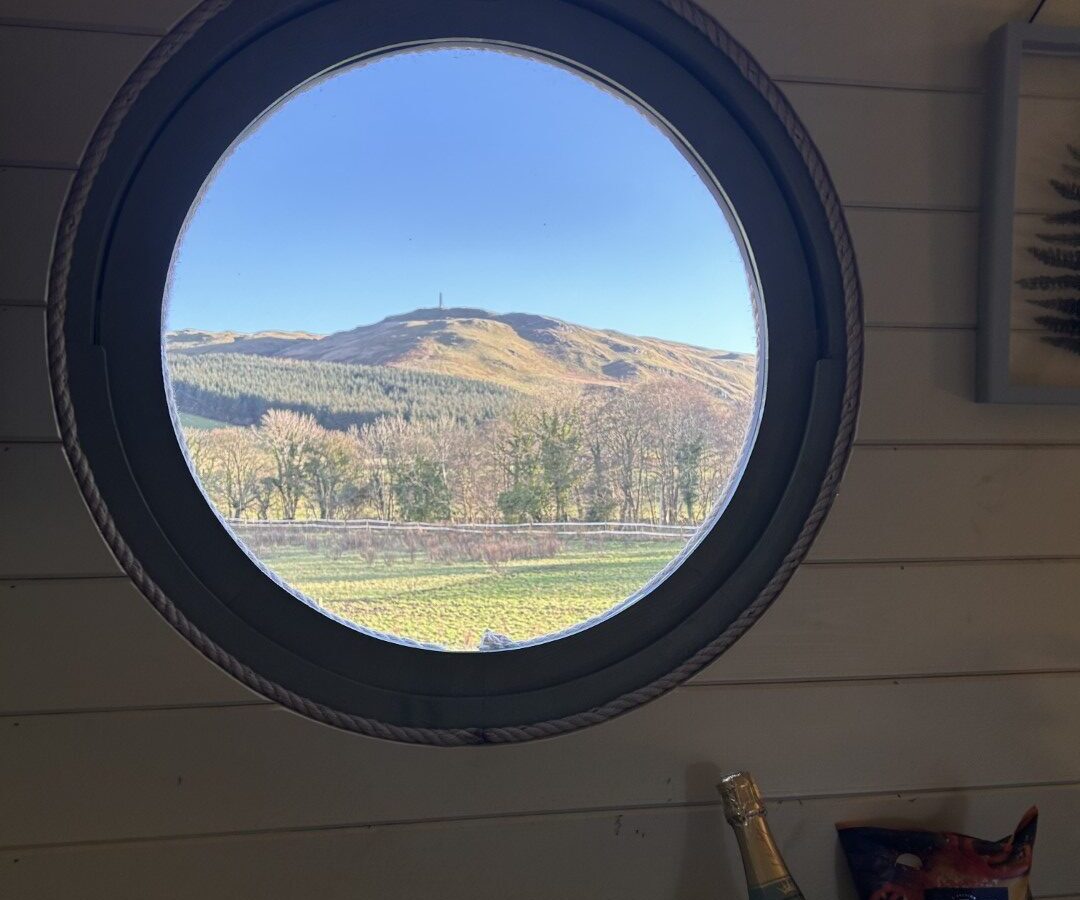 A circular porthole window in a wooden wall frames a scenic view of the hilly landscape at Freedom Fields, with a clear blue sky. Below the window, there's a wooden shelf holding a bottle of wine, a bag of snacks, and a chocolate bar—perfect for glamping in nature's embrace.