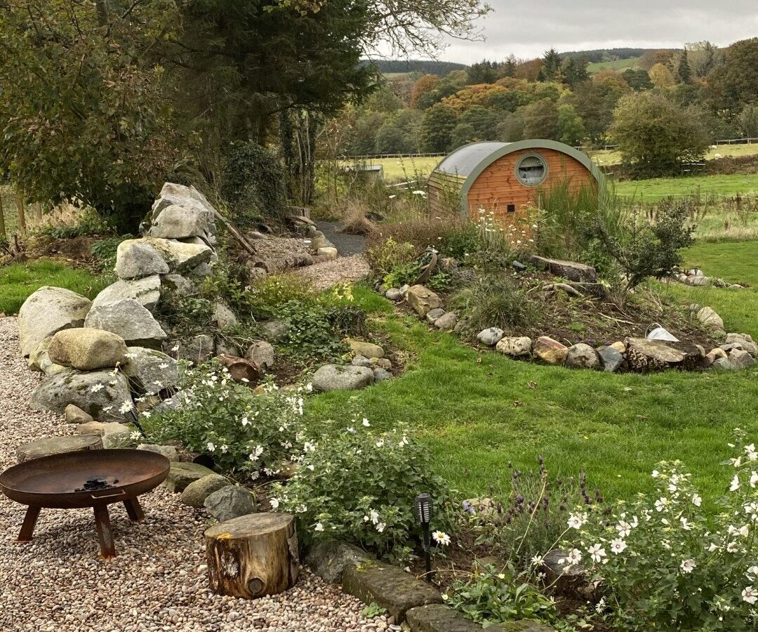 A tranquil garden scene with a gravel path, surrounded by lush greenery and white flowers. There is a round wooden cabin with a curved roof in the background, perfect for glamping enthusiasts at Freedom Fields. In the foreground, a fire pit and tree stump seat invite relaxation, with hills and trees in the distance.