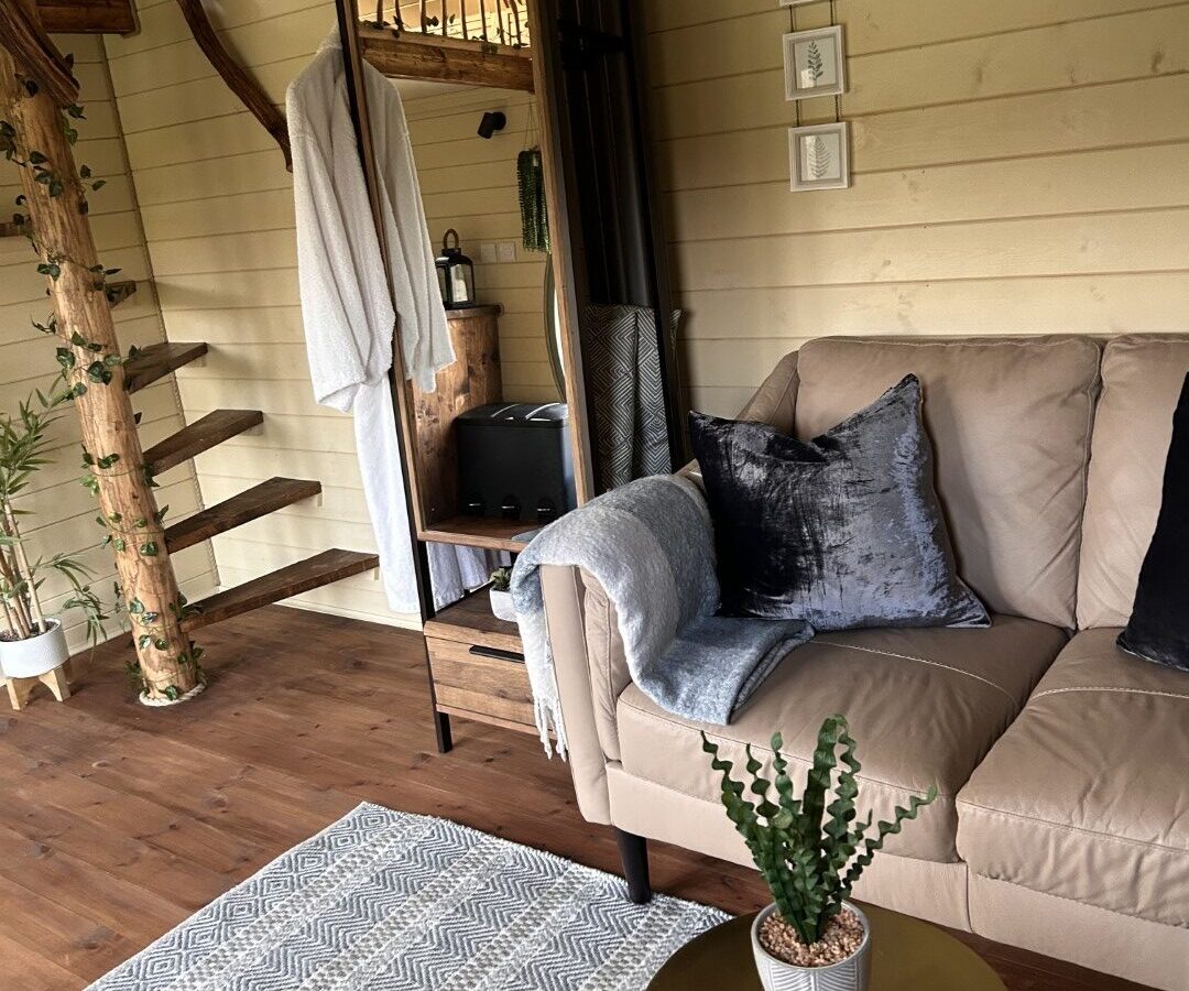 A cozy interior at Freedom Fields features a beige couch with gray pillows and a throw blanket. A small round table with a potted plant sits nearby. Wooden stairs with a rustic touch lead to an upper level. Light wood walls are adorned with small framed pictures and shelves, perfect for glamping comfort.