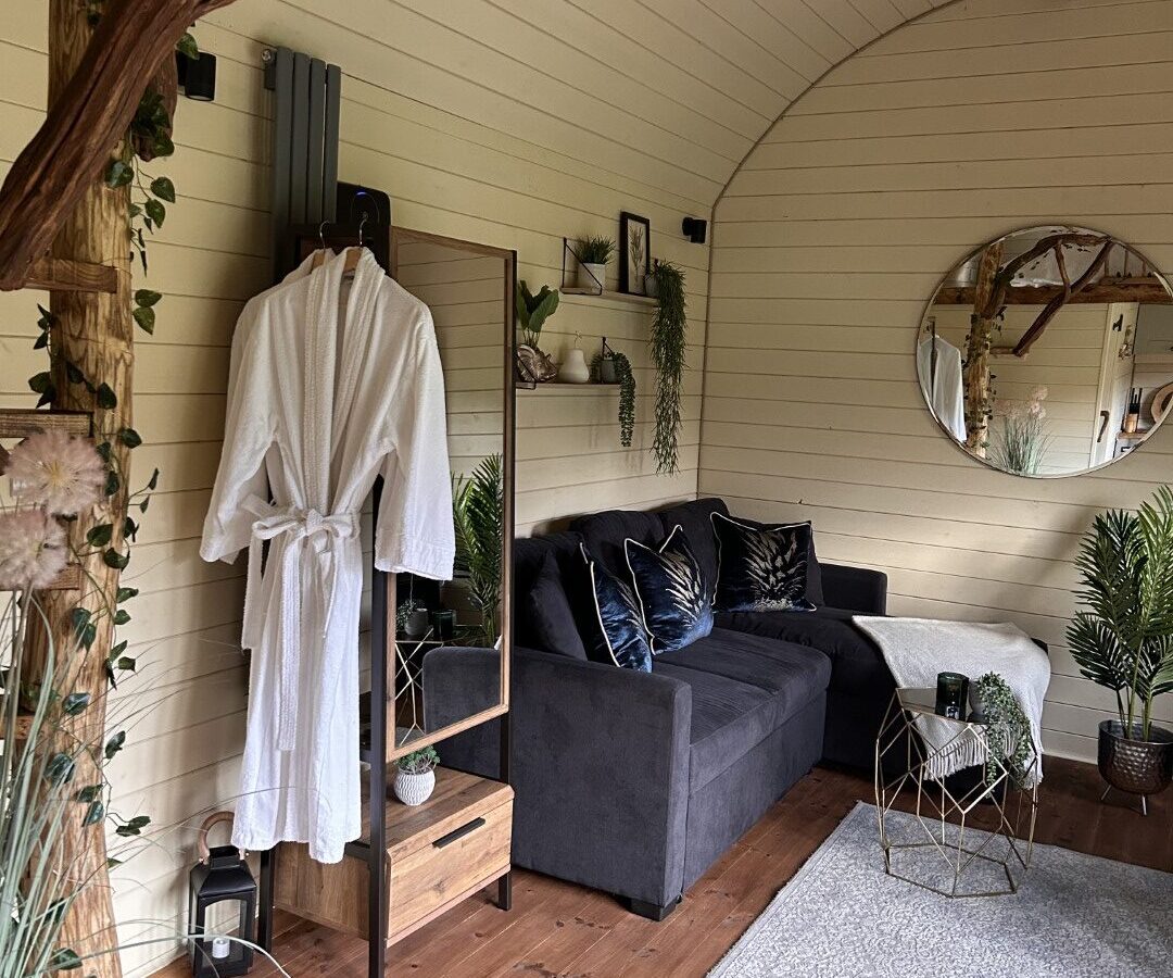 A cozy spa room at Freedom Fields Glamping boasts a wooden interior with a dark gray sofa adorned with cushions, a large round mirror, and plants on the walls. A white bathrobe hangs on a stand beside a full-length mirror, while exposed wooden beams overhead are decorated with hanging lights.