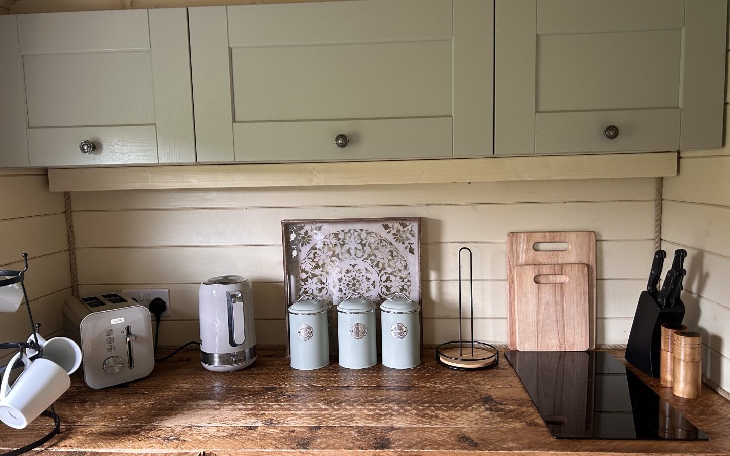 A kitchen countertop featuring a coffee maker, toaster, tea kettle, three matching canisters, and a decorative cutting board against the back wall. A knife block and two cutting boards are to the right, with light sage overhead cabinets that evoke a sense of freedom like open fields.