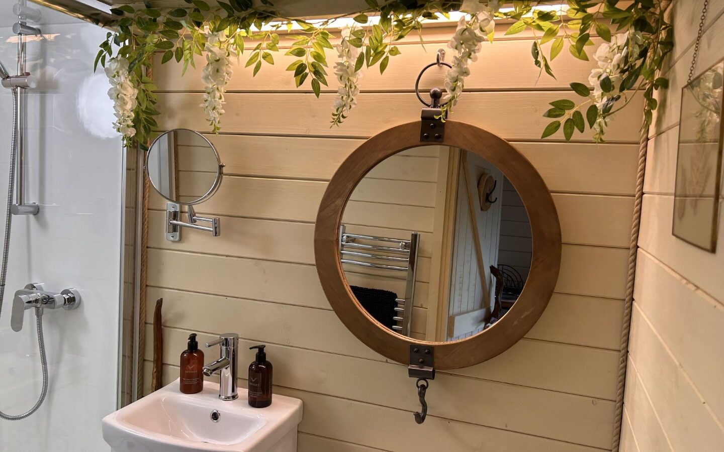 A cozy bathroom, perfect for glamping, features a round wooden-framed mirror hanging on a beige paneled wall, with a small sink below it. Two brown soap dispensers sit on the sink. Above the mirror, white flowers and green leaves are draped. A shower area with a glass door is to the left.