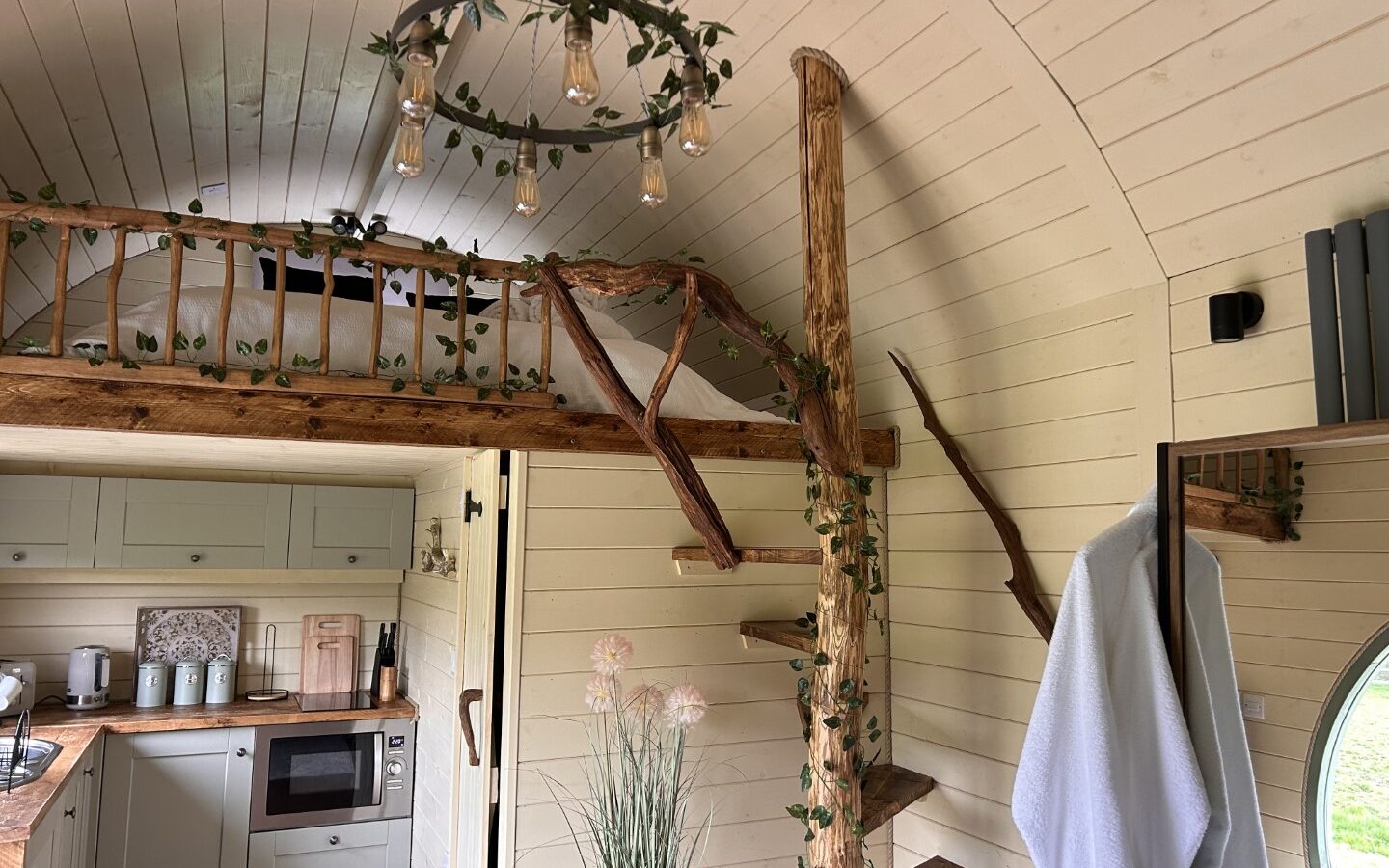 A cozy tiny home interior with wood-paneled walls at Freedom Fields Glamping. A rustic, vine-adorned staircase leads to a loft. The kitchenette features a microwave, stove, and sink. A round window and hanging lamp with vines add charm. Towels hang by the doorway, and decorative plants sit on a table.