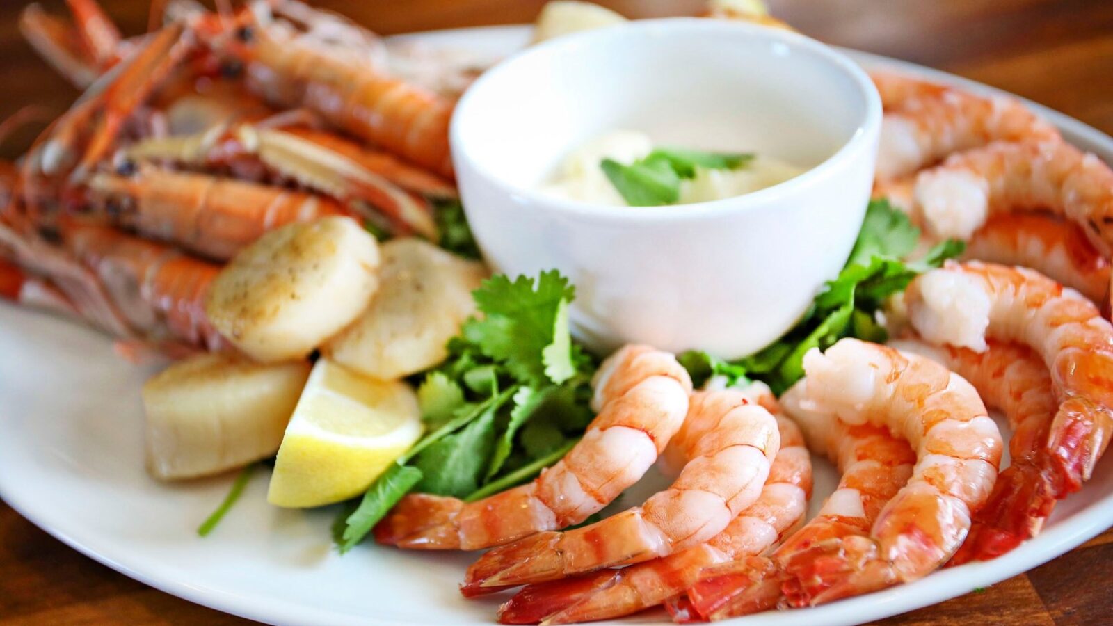 A plate of seafood features peeled shrimp, scallops, and langoustines arranged around a small bowl of creamy white sauce. Garnished with lemon wedges and fresh cilantro, this vibrant dish is set on a wooden surface, reminiscent of a serene cruise through Argyll's coastal waters.