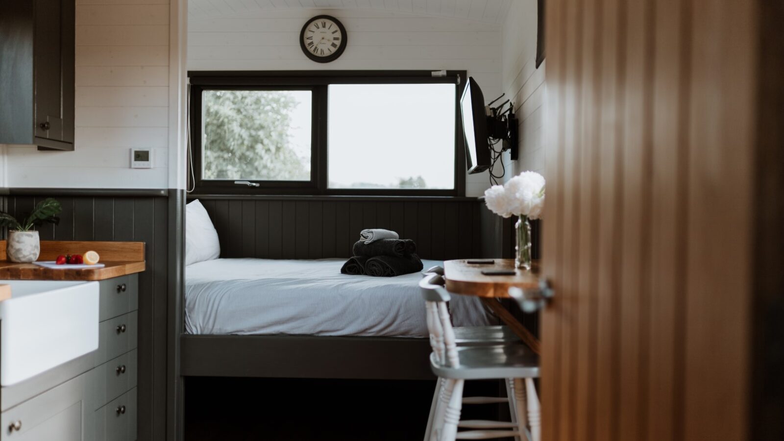 Cozy room in the Ginger Nut Shepherds Hut, featuring a bed under a window, a wooden desk with a chair, and a clock above. White flowers in a vase add charm to the desk.