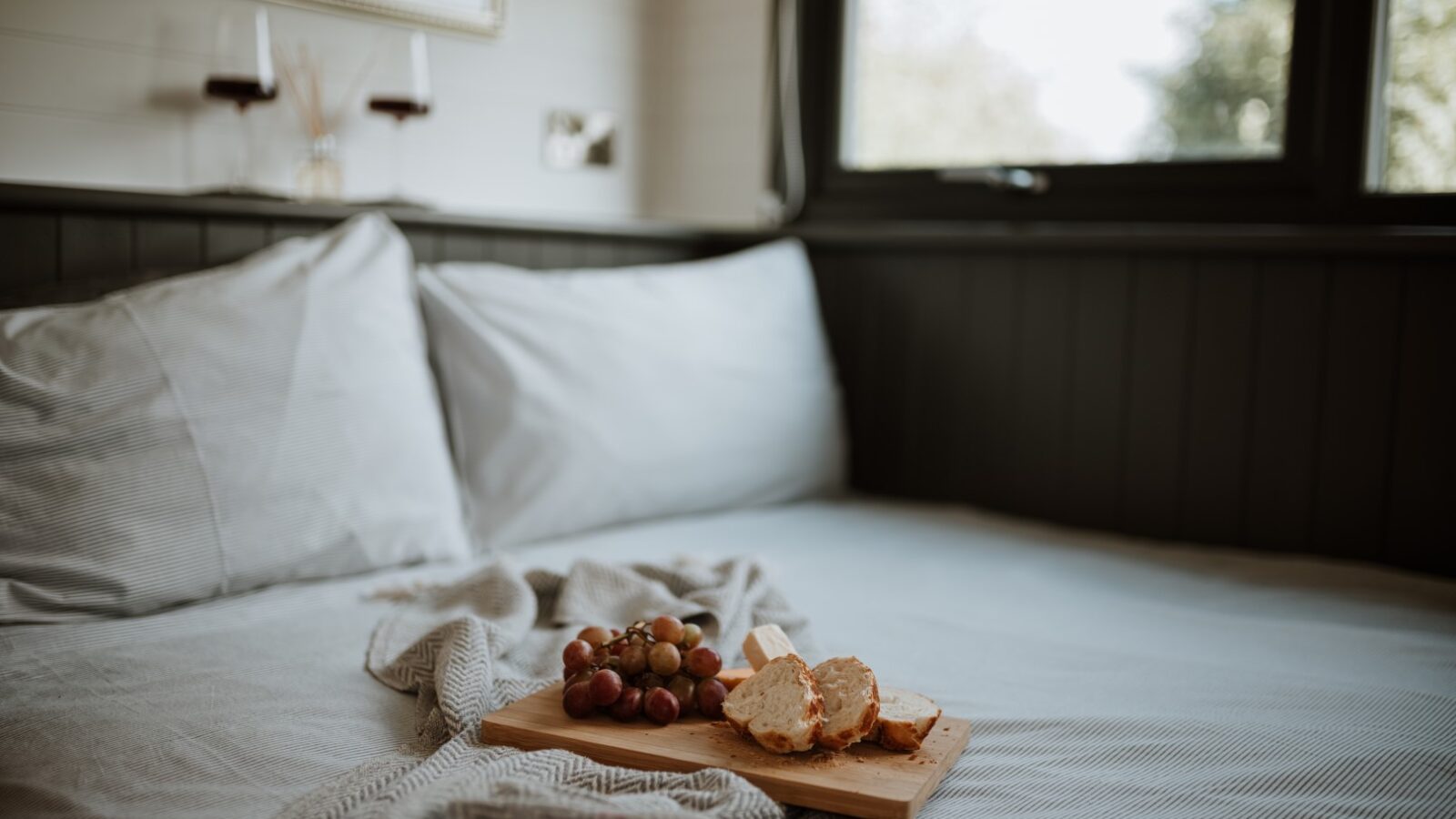 A wooden tray with bread, cheese, and grapes rests on a pillow-laden bed in a cozy shepherd's hut, offering a delightful view of nature through the window.