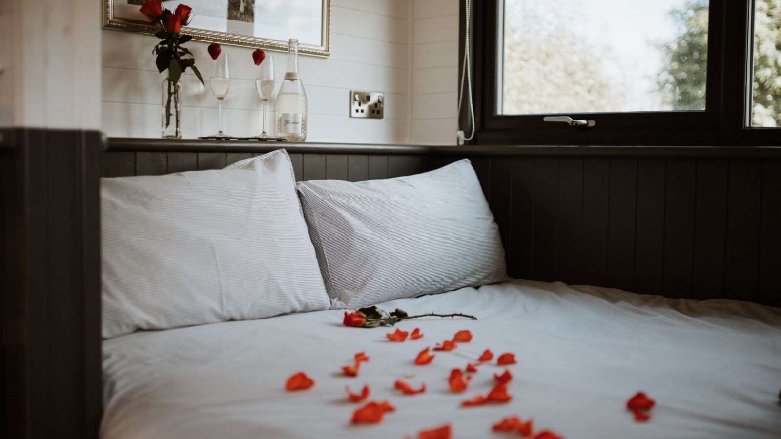 A cozy shepherd's hut features a bed with white pillows and sheets, scattered red rose petals, wine glasses, a rose in a vase, and a window with blinds.