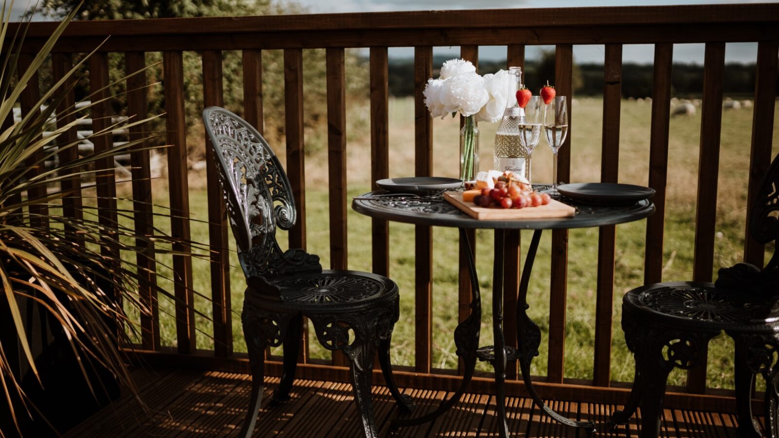 Two ornate chairs and a round table on the wooden terrace of Ginger Nut Shepherds Hut, adorned with flowers, glasses, and a platter of fruits. A serene grass field stretches into the background.