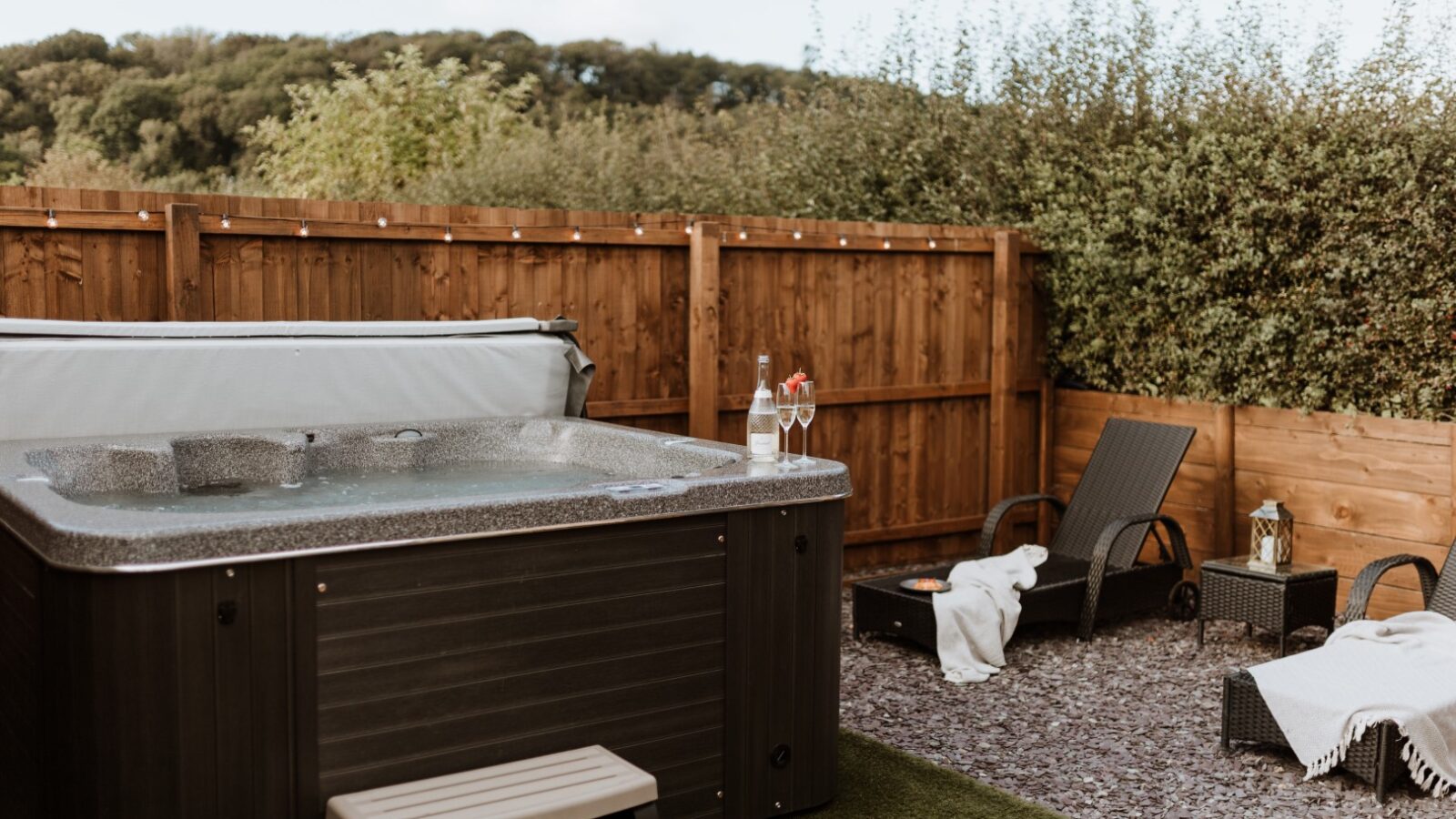 The Ginger Nut Shepherds Hut features an outdoor hot tub with a cover, nestled among lounge chairs and towels on a tranquil gravel patio, set against a rustic wooden fence and lush greenery.