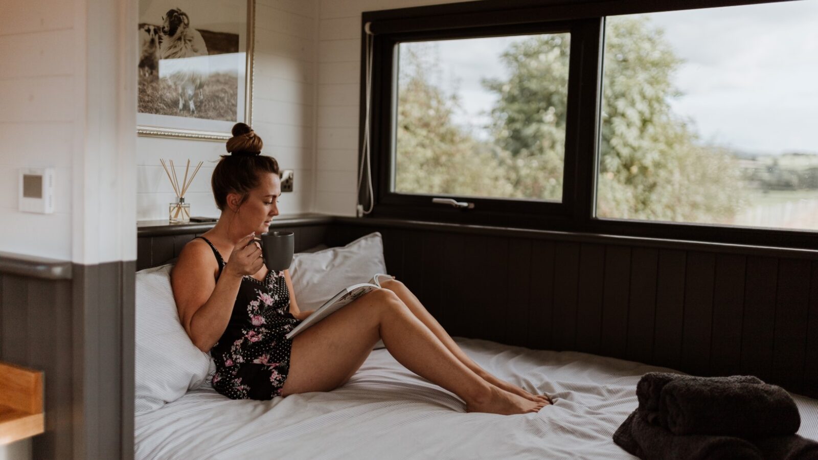 In the cozy embrace of her Ginger Nut Shepherds Hut, a woman in floral pajamas sips from a mug and immerses herself in a book, while the large window reveals lush greenery outside.