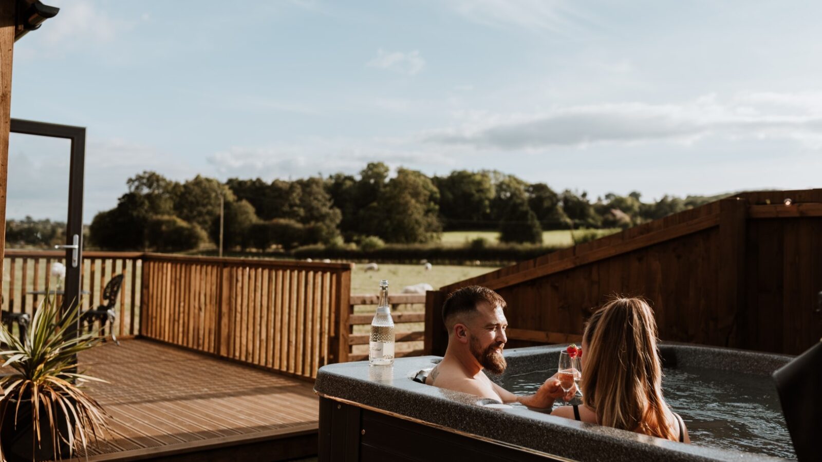 A couple unwinds in an outdoor hot tub on a wooden deck beside a cozy shepherd's hut, surrounded by countryside views and a bottle of drink.