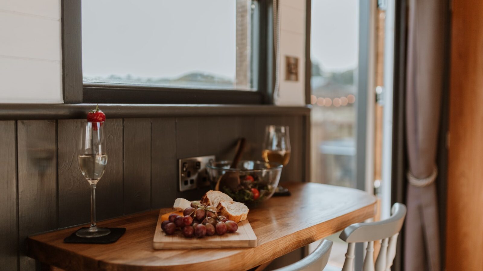 The cozy Ginger Nut Shepherds Hut features a charming wooden kitchen counter adorned with grapes, bread, a bowl of salad, and two wine glasses—one elegantly garnished with red—perfectly positioned near the window.