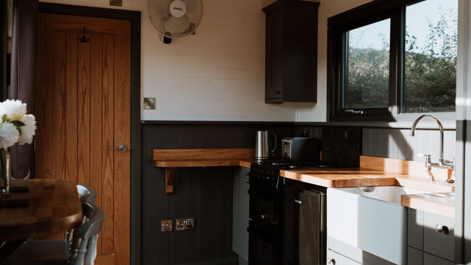 Compact kitchen with wood accents in the Ginger Nut Shepherds Hut, featuring an oven, sink, and wooden countertops. A fan is mounted on the wall.