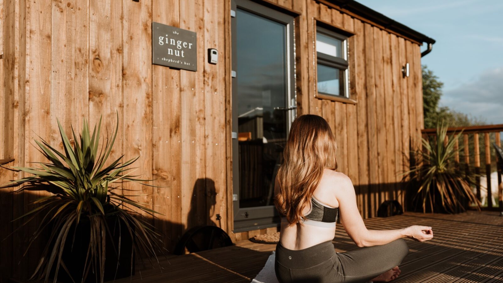 In front of the rustic Ginger Nut Shepherds Hut, a woman in activewear gracefully practices yoga on her mat, surrounded by lush potted plants.