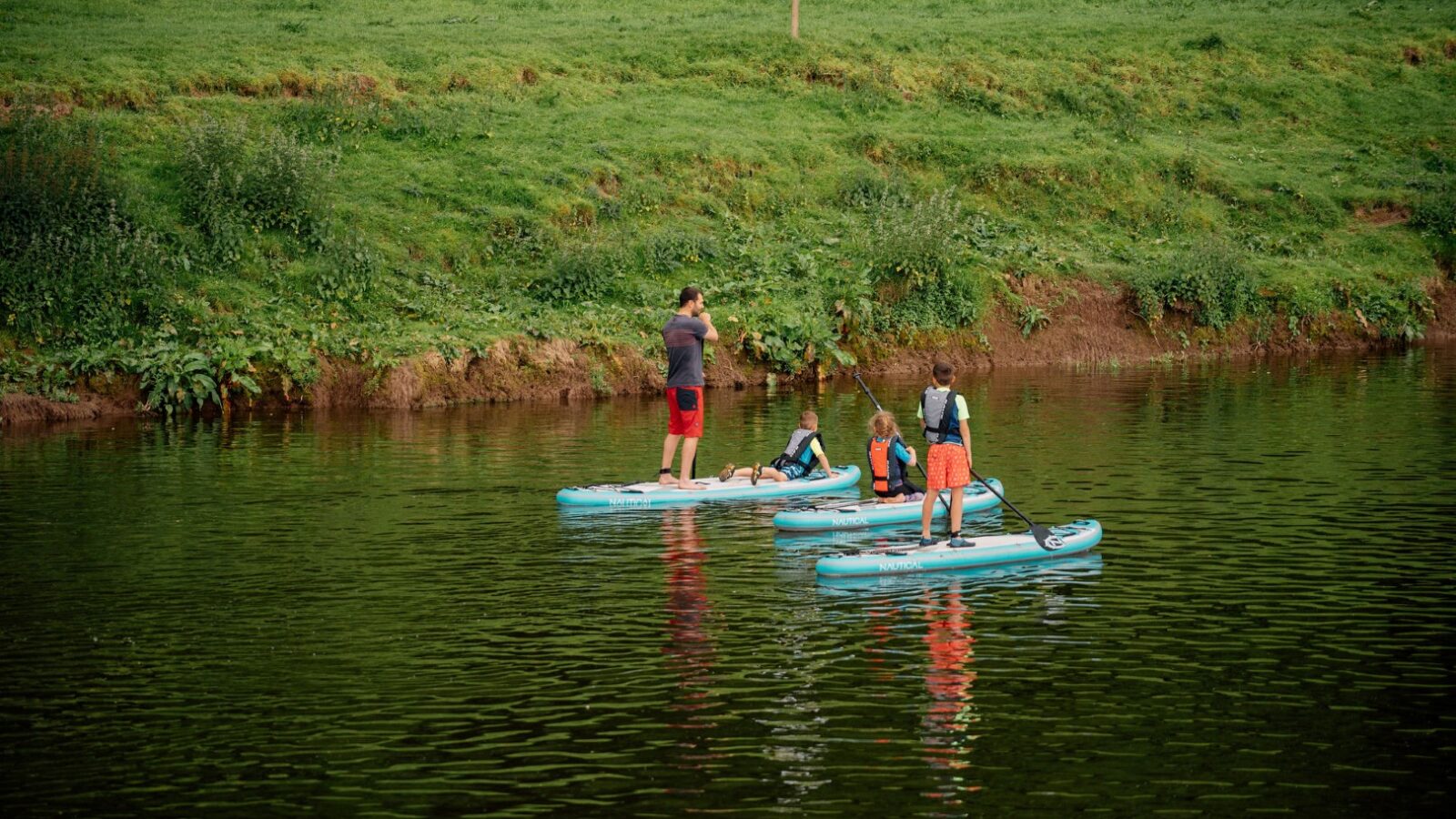 Three people stand on blue paddleboards in a calm river, surrounded by the serenity of nature near a grassy bank. One person holds a paddle upright while the others sit. A sign is visible on the green bank in the background, hinting at nearby glamping adventures.