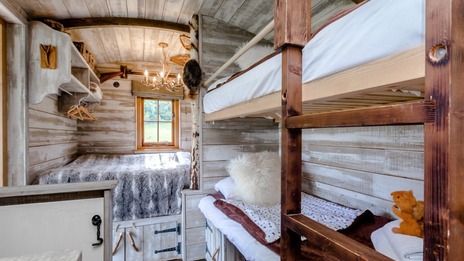 A cozy, rustic bedroom in a ranch cabin features wooden walls and ceiling with a soft chandelier. The room includes bunk beds and a larger bed, both adorned with fluffy bedding. A small window lets in natural light, and there's a teddy bear on the bunk bed, perfect for those seeking loose reins from daily hustle.