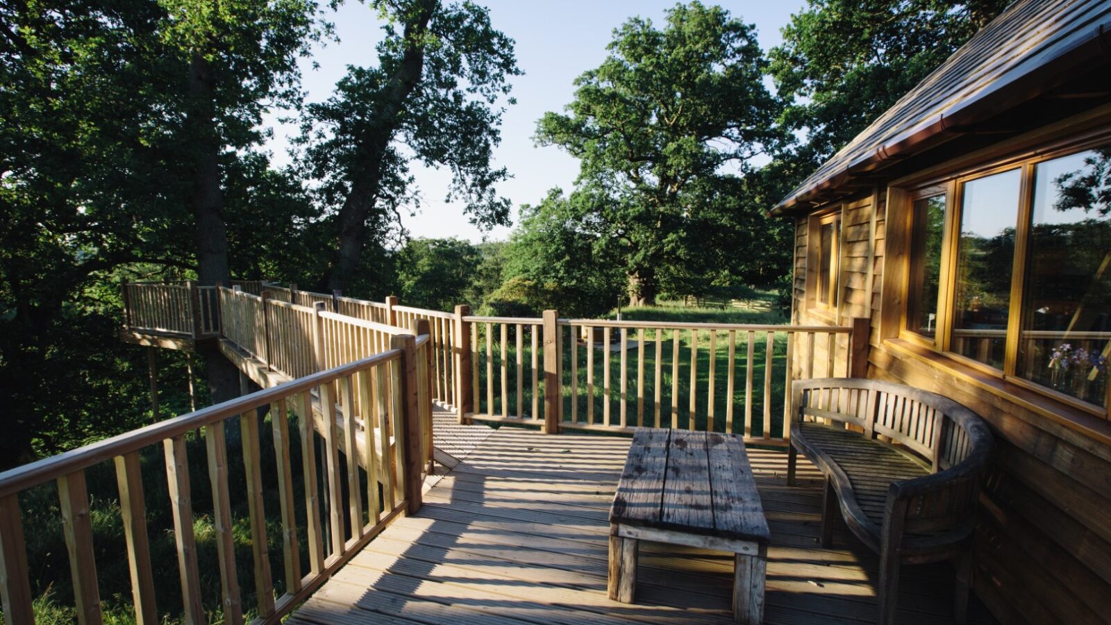 A wooden deck extends from the Netherby Treehouse, a perfect nature retreat surrounded by lush, green trees. The deck features a rustic wooden bench and a matching table, inviting relaxation amidst the peaceful setting. Sunlight filters through the leaves, casting gentle shadows on this charming treehouse rental.