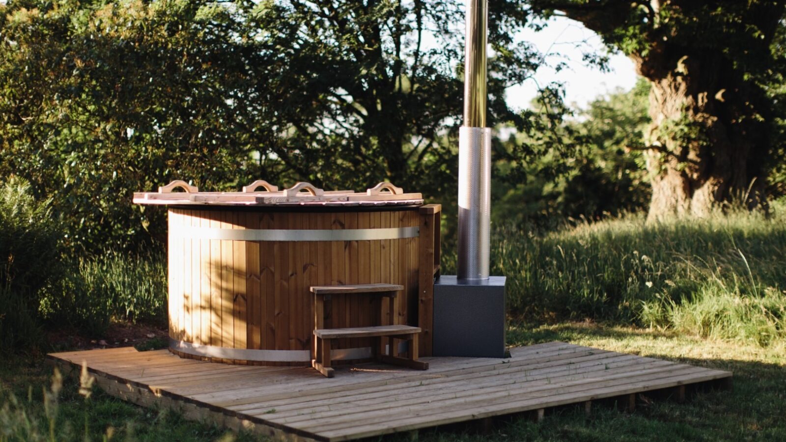 A wooden hot tub surrounded by grass and trees is placed on a raised wooden deck at the Netherby Treehouse. The tub has a metal chimney for heating and wooden steps leading up to it. The scene is bathed in natural daylight, suggesting a serene, secluded outdoor setting.