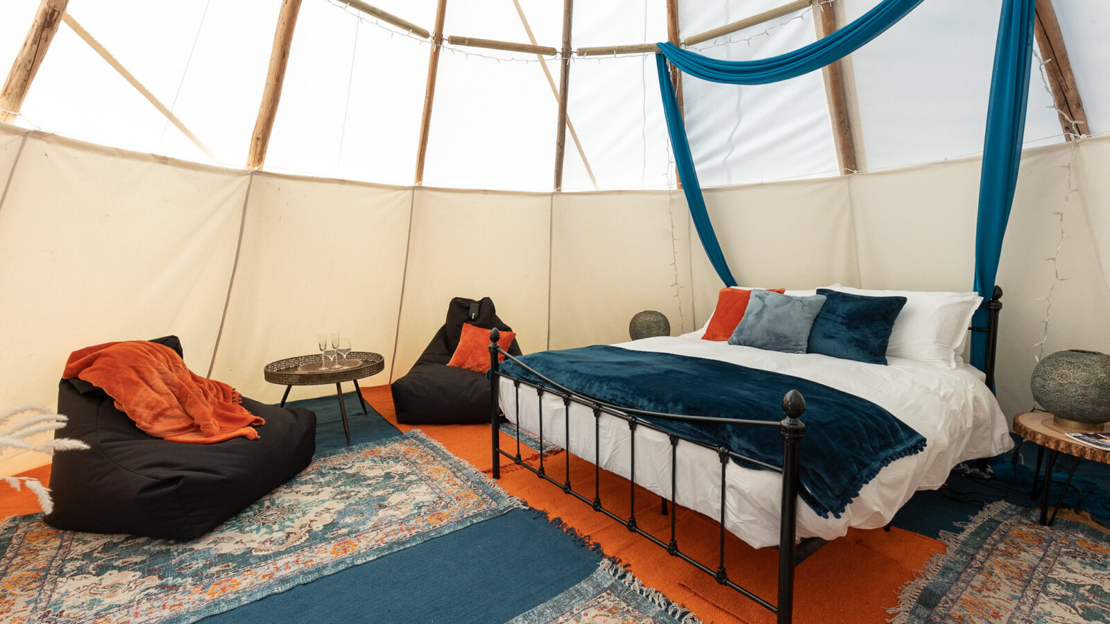 Interior of a cozy yurt at the HARTA retreat, decorated with blue and orange accents. Features a black metal bed with matching pillows, black bean bag chairs, a small wooden side table with a globe lamp, patterned rugs, and blue drapes hanging in the background.