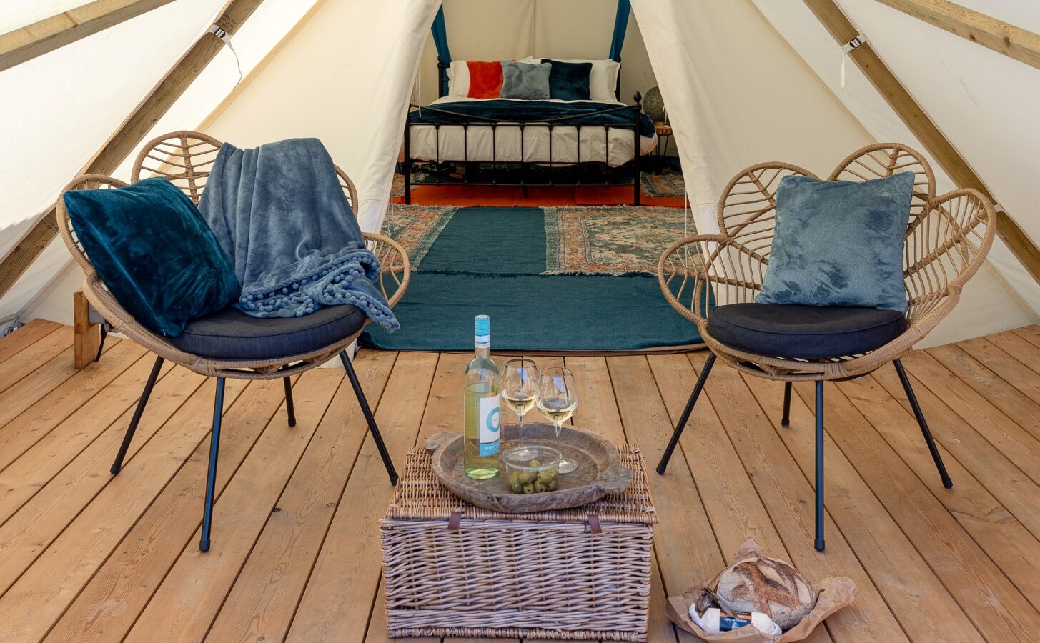 A HARTA glamping tent interior featuring two wicker chairs with dark cushions and plush throws, a wicker table with a bottle of wine, two glasses, and snacks. In the background, a cozy bed with plush bedding sits on a patterned rug. The wooden floor adds to the perfect retreat ambiance.