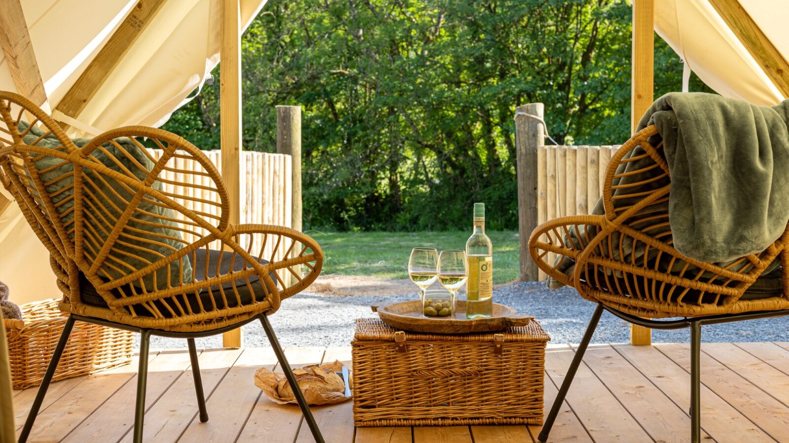 A cozy outdoor glamping retreat featuring two wicker chairs with cushions, a wicker table with a bottle of white wine, two filled glasses, and a snack. The scene is under a tent canopy branded with HARTA, facing a lush green background on a sunny day with a serene atmosphere.