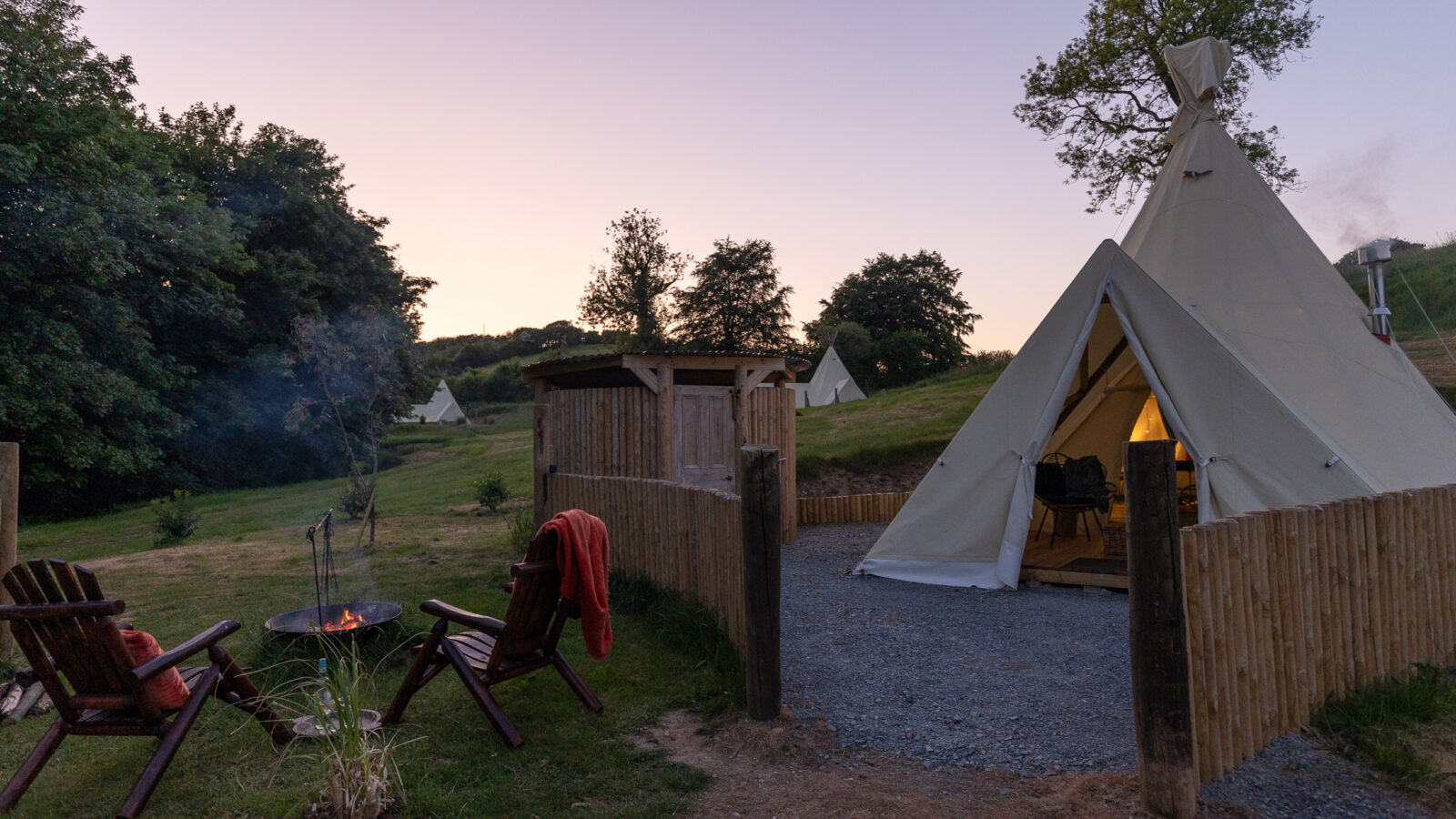 The image shows a serene HARTA retreat at dusk with a cozy tipi tent illuminated from within. In front of the tent, there are two wooden chairs and a fire pit. The campsite is surrounded by green trees and hills. The sky is a soft gradient of twilight colors.