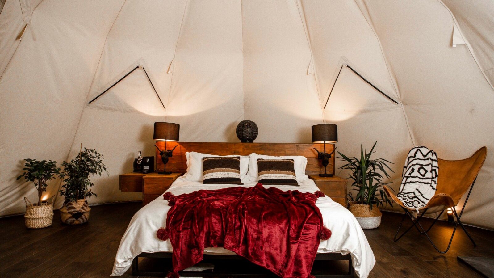 A cozy bedroom setup inside a tent features a wooden bed with white sheets and a red throw blanket. The retreat is accented with plants, two side tables with lamps, and a patterned chair. Soft lighting and rustic decor elements create an inviting atmosphere at HARTA.