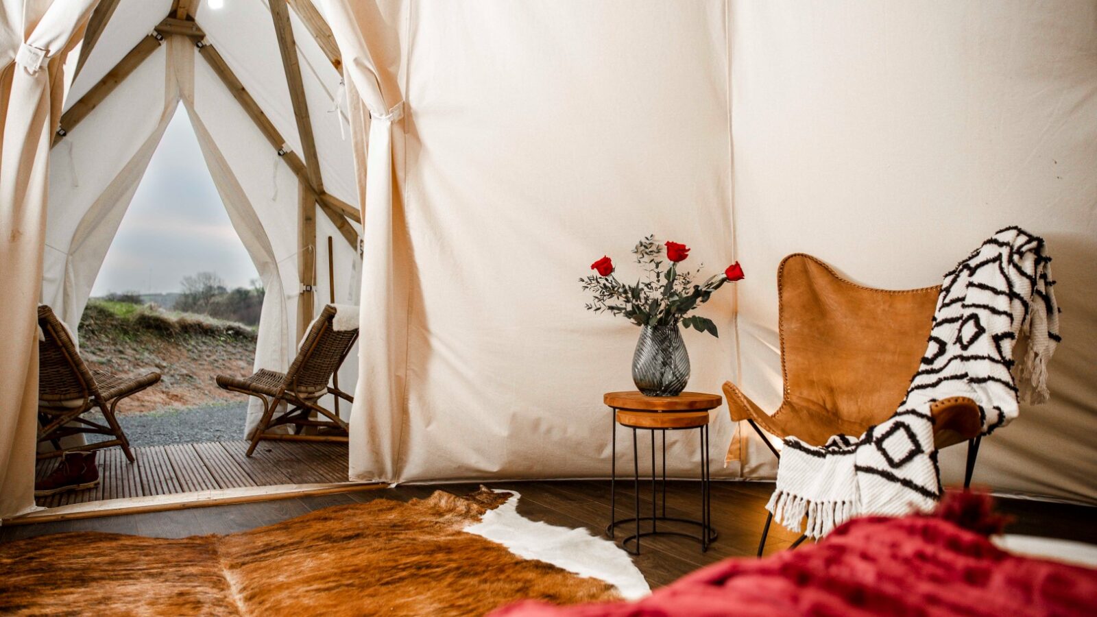 A cozy glamping tent interior with a leather armchair draped with a black and white blanket, a small table with a vase of red roses, and a cowhide rug. The scene is perfect for an HARTA retreat, complete with two outdoor chairs positioned outside the tent facing the scenic view.