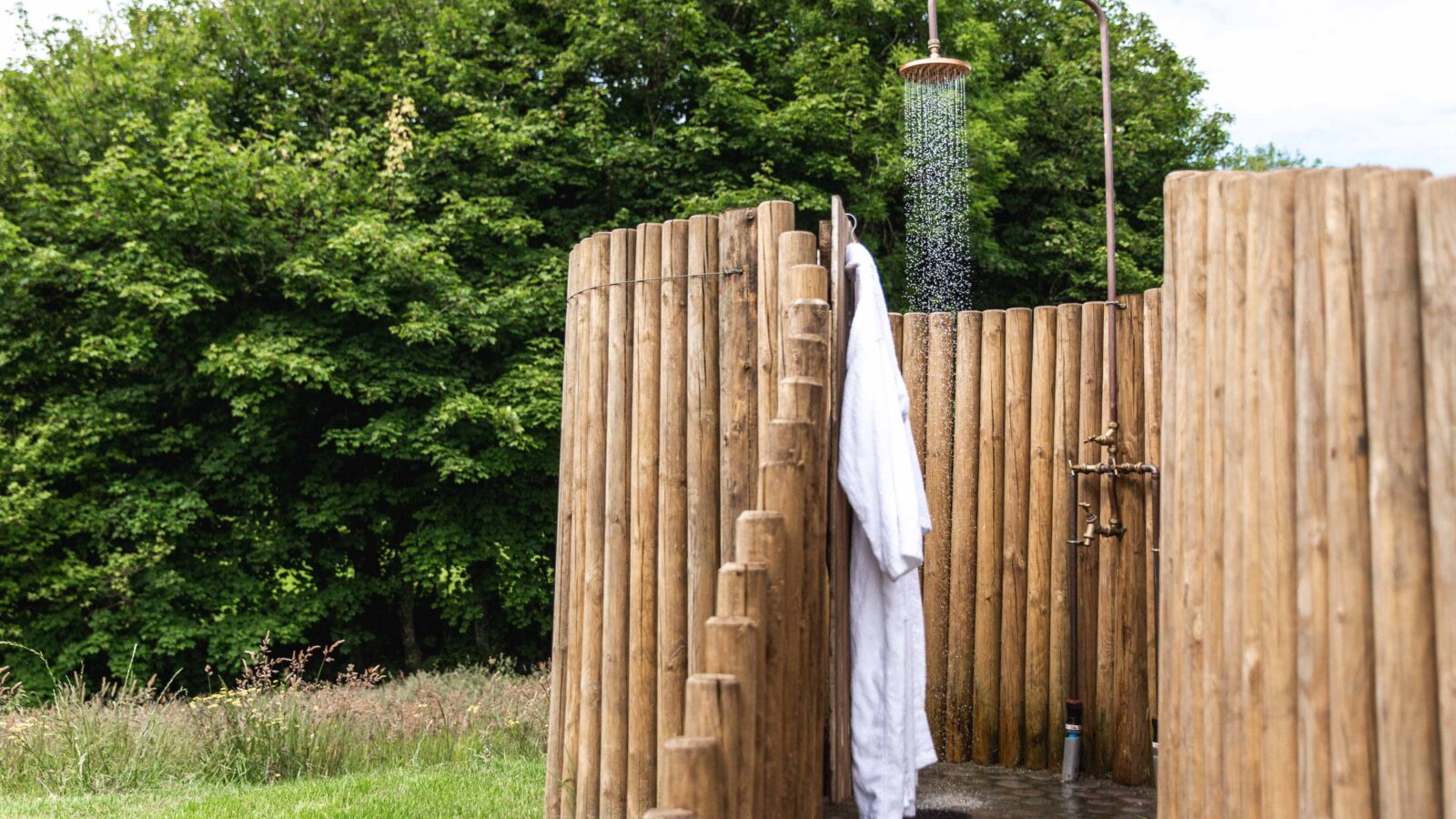 An outdoor shower with wooden walls and a metal showerhead pouring water stands in a grassy area with trees in the background. A white robe hangs on the wooden partition. The rustic setting exudes tranquility, making it an ideal HARTA retreat surrounded by nature.