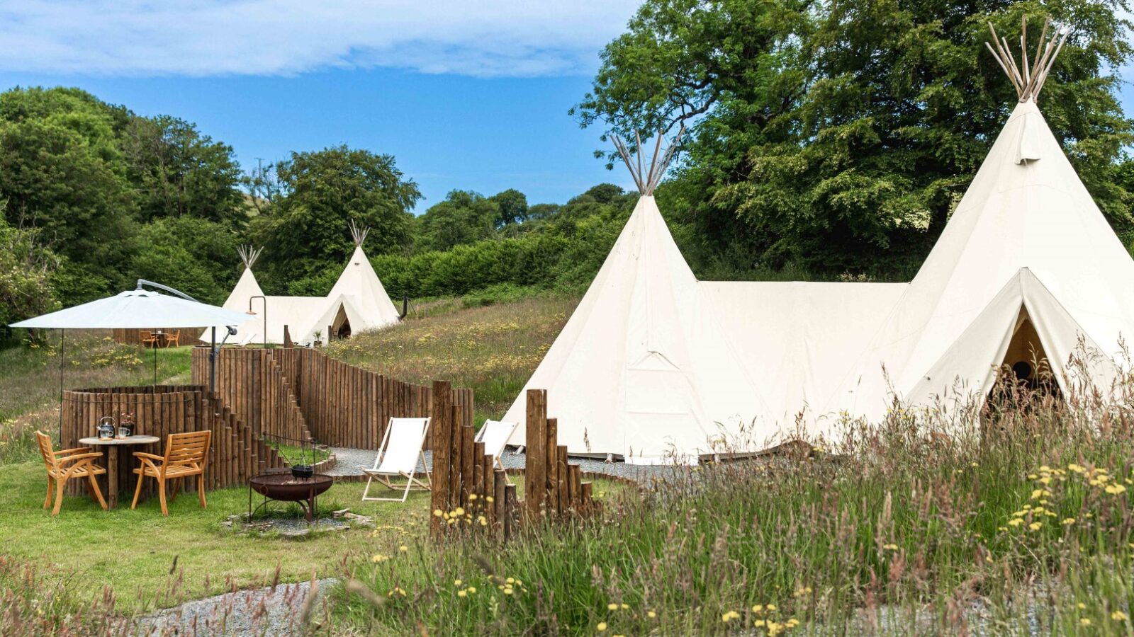 A scenic outdoor glamping retreat, HARTA, features large white teepee tents surrounded by lush greenery and wildflowers. A wooden table with chairs and a large umbrella invite relaxation by the fire pit in the foreground under a clear blue sky.