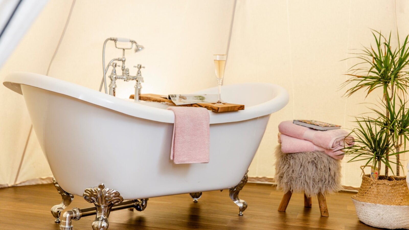 A white clawfoot bathtub is set up in an elegantly decorated space with a wooden floor. A pink towel hangs over the tub, and a wooden tray holding a glass of champagne rests on the edges. Next to the tub is a small stool with pink towels and a potted plant, creating the perfect retreat for relaxation.