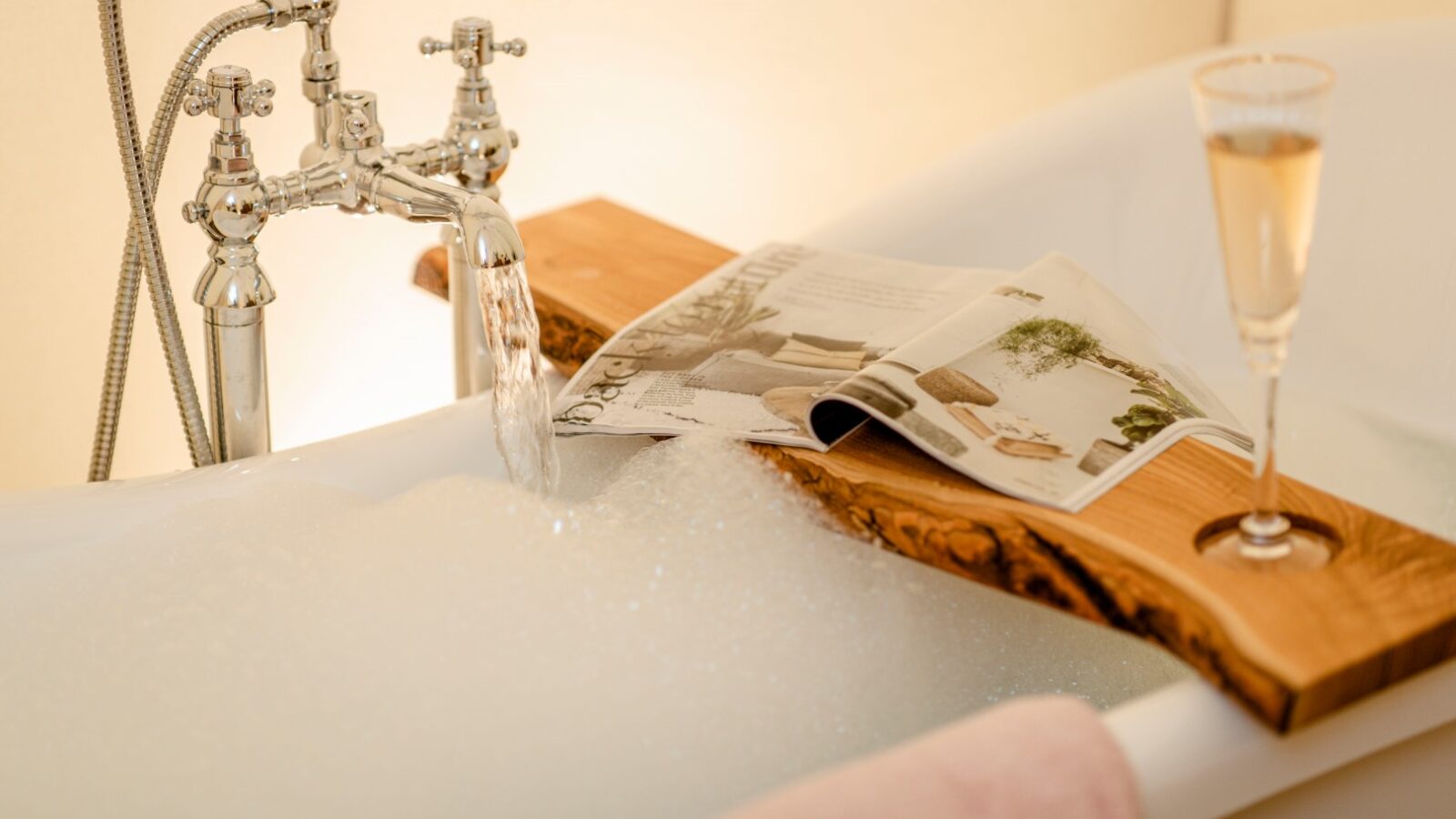 A luxurious bubble bath with running water in a white bathtub sets the scene for a tranquil retreat. A wooden tray rests on the tub's edges, holding an open magazine and a glass of HARTA champagne. A towel hangs over the tub's side, completing the relaxing atmosphere.