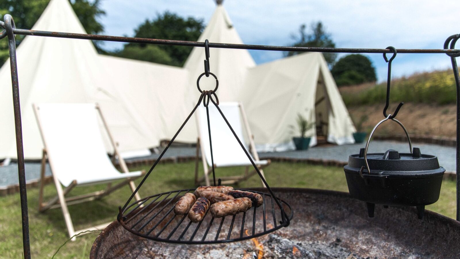 A rustic outdoor scene at a HARTA retreat features a barbecue grill with sausages cooking over an open flame. Nearby, a black cast iron pot hangs over the fire. In the background, there are two white canvas tents and two empty deck chairs set up on a grassy area with trees.