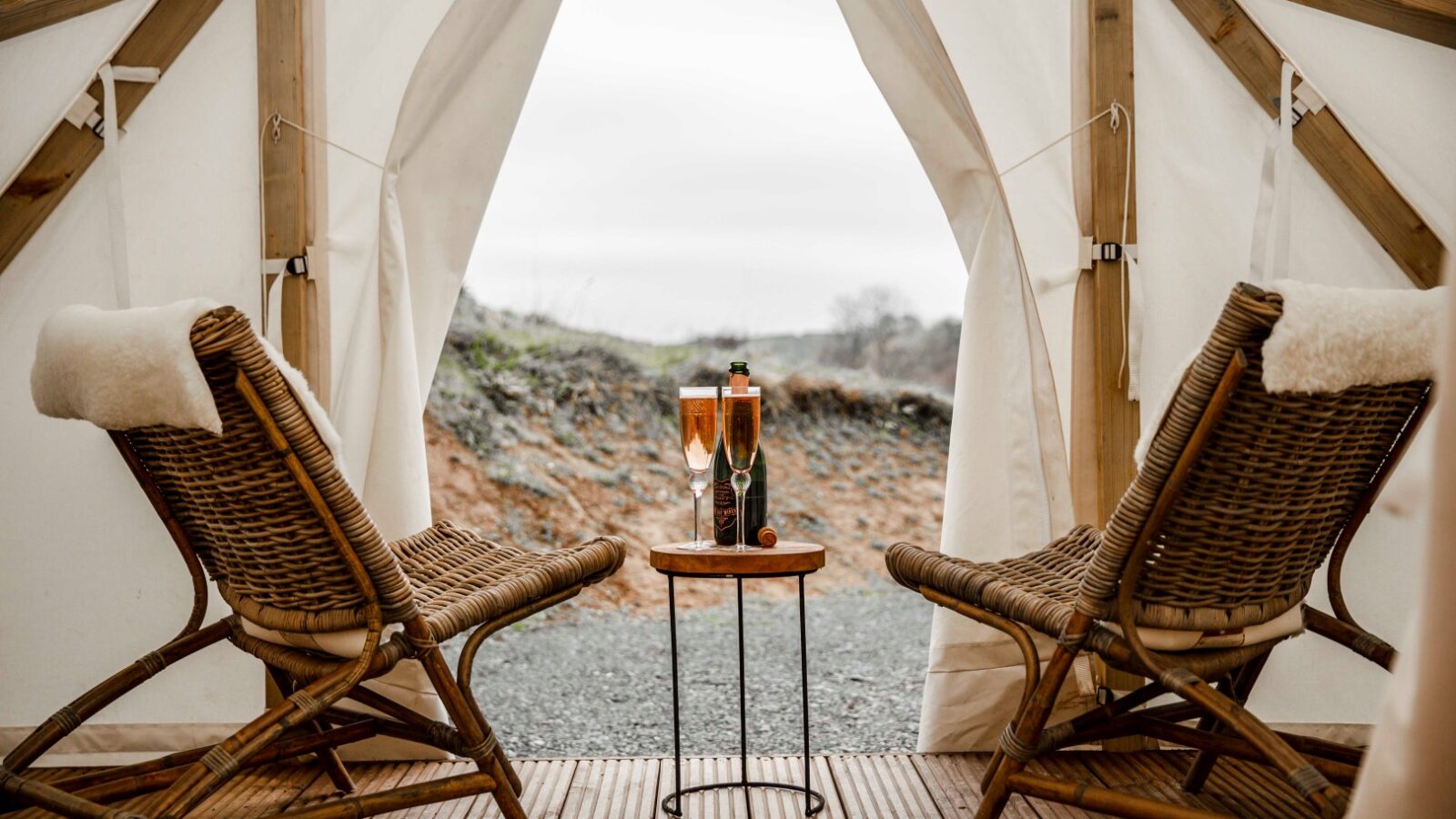 A cozy glamping tent with wooden frames and a wooden floor features two wicker armchairs with furry seat covers. A small wooden table between the chairs holds two filled champagne flutes and a champagne bottle, offering the perfect retreat overlooking a view of a gravel path and greenery.