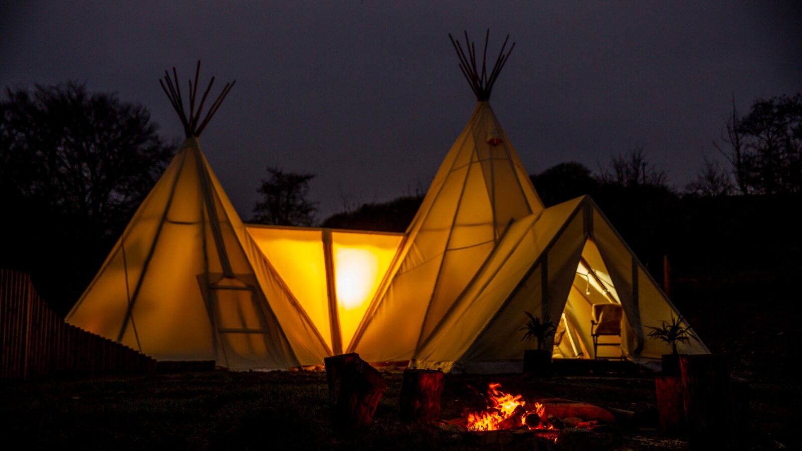 A nighttime retreat features two illuminated HARTA tents with wooden supports, shaped like teepees. The tents glow warmly against the dark sky. In the foreground, there's a small, crackling campfire with several tree stumps arranged around it.