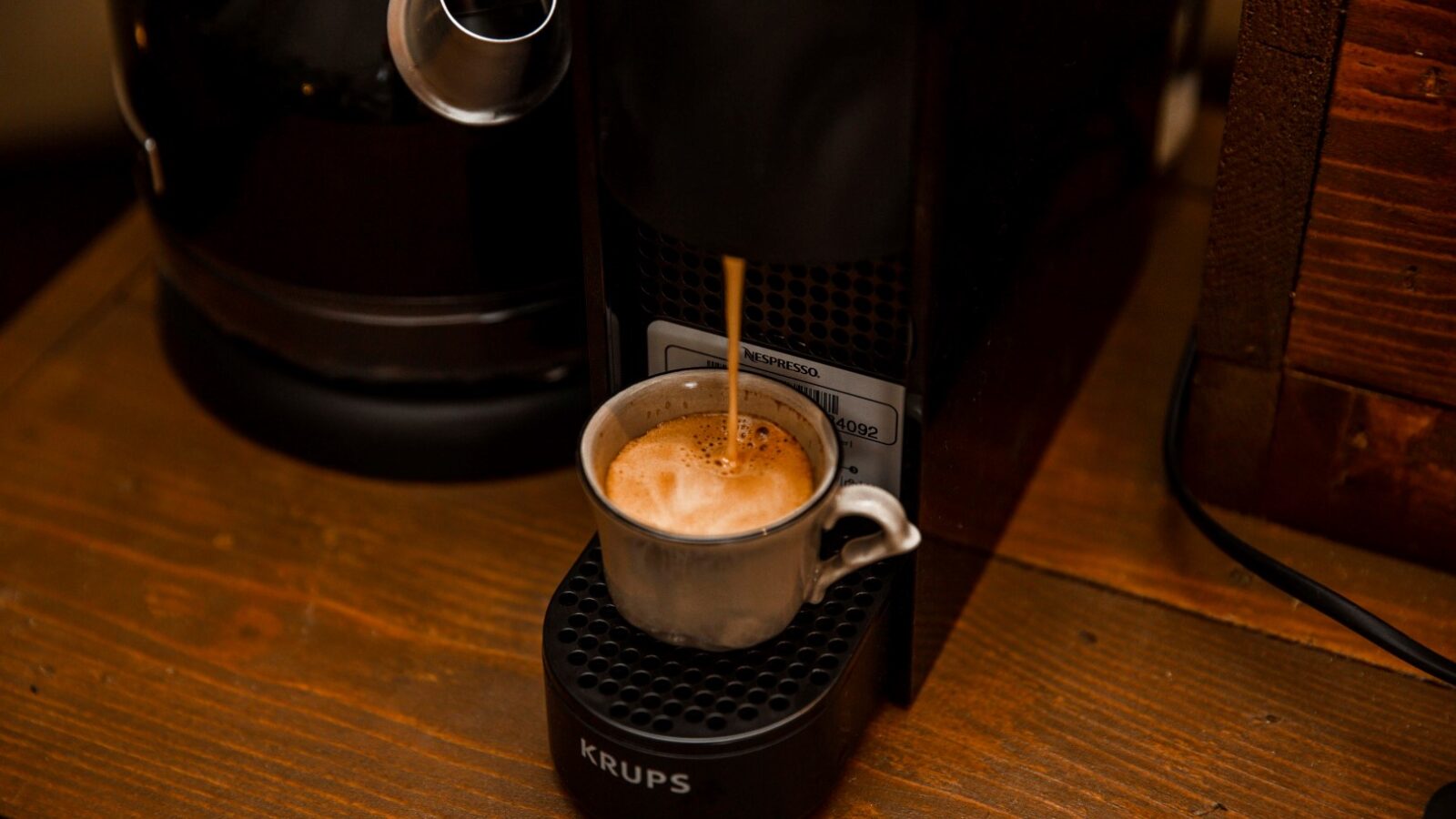 A small HARTA espresso machine dispenses coffee into a silver and white ceramic cup placed on its platform. The scene is set on a wooden surface, reminiscent of a cozy retreat, with another coffee-related appliance partially visible in the background.