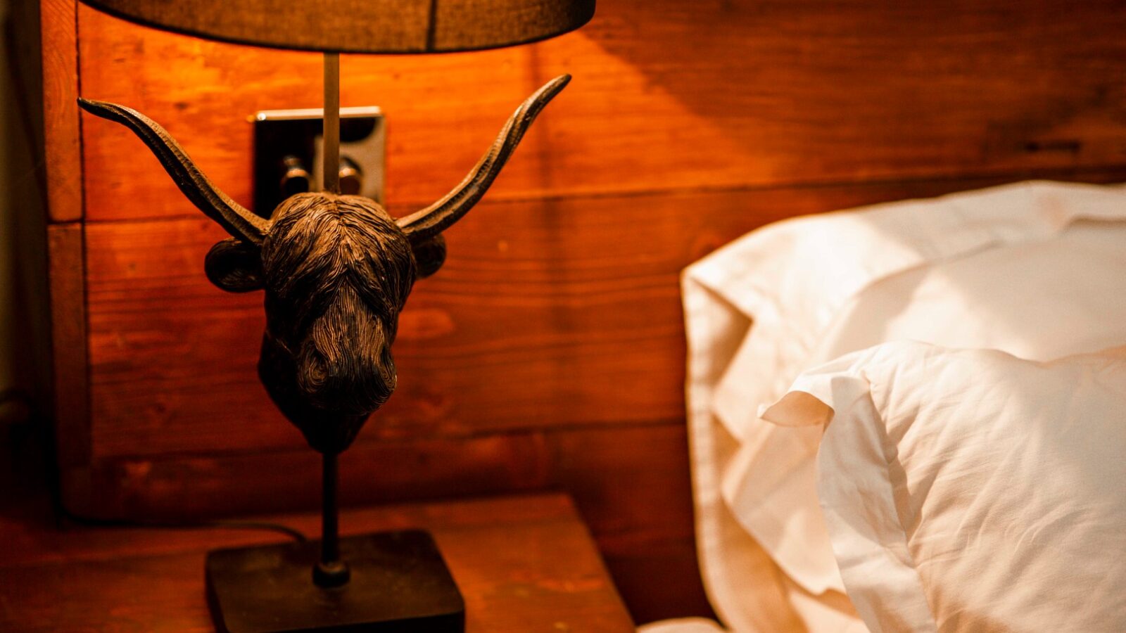 A dimly lit rustic bedroom retreat features a unique bedside lamp with a bull's head base and a brown lampshade. The HARTA lamp rests on a wooden nightstand next to a bed with white pillows and linens, set against a wooden plank wall.