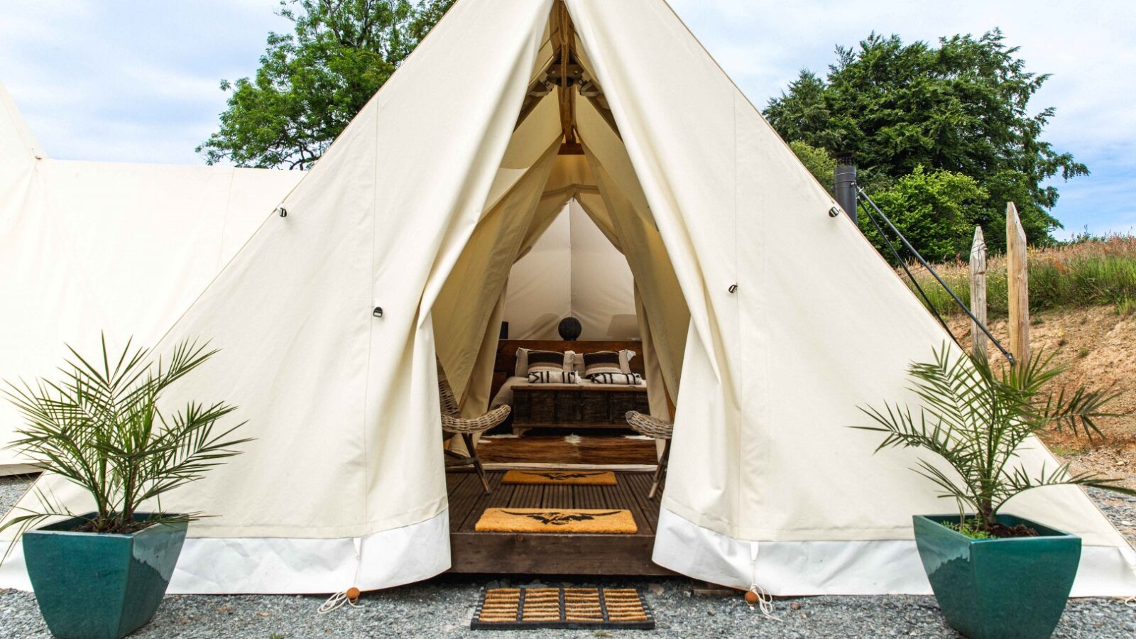 A spacious cream-colored tent set up outdoors with its entrance flap open beckons you to a serene retreat. Inside, a cozy arrangement of wooden furniture, including chairs and a small table from HARTA, invites relaxation. Two green potted plants flank the tent entrance, with trees in the background enhancing the tranquility.