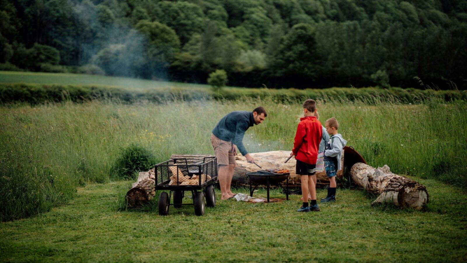 In a lush, green field perfect for glamping, a man is grilling food over a fire, accompanied by two children. A small cart with logs nestles nearby, framed by trees in the background. Smoke from the fire drifts upward.